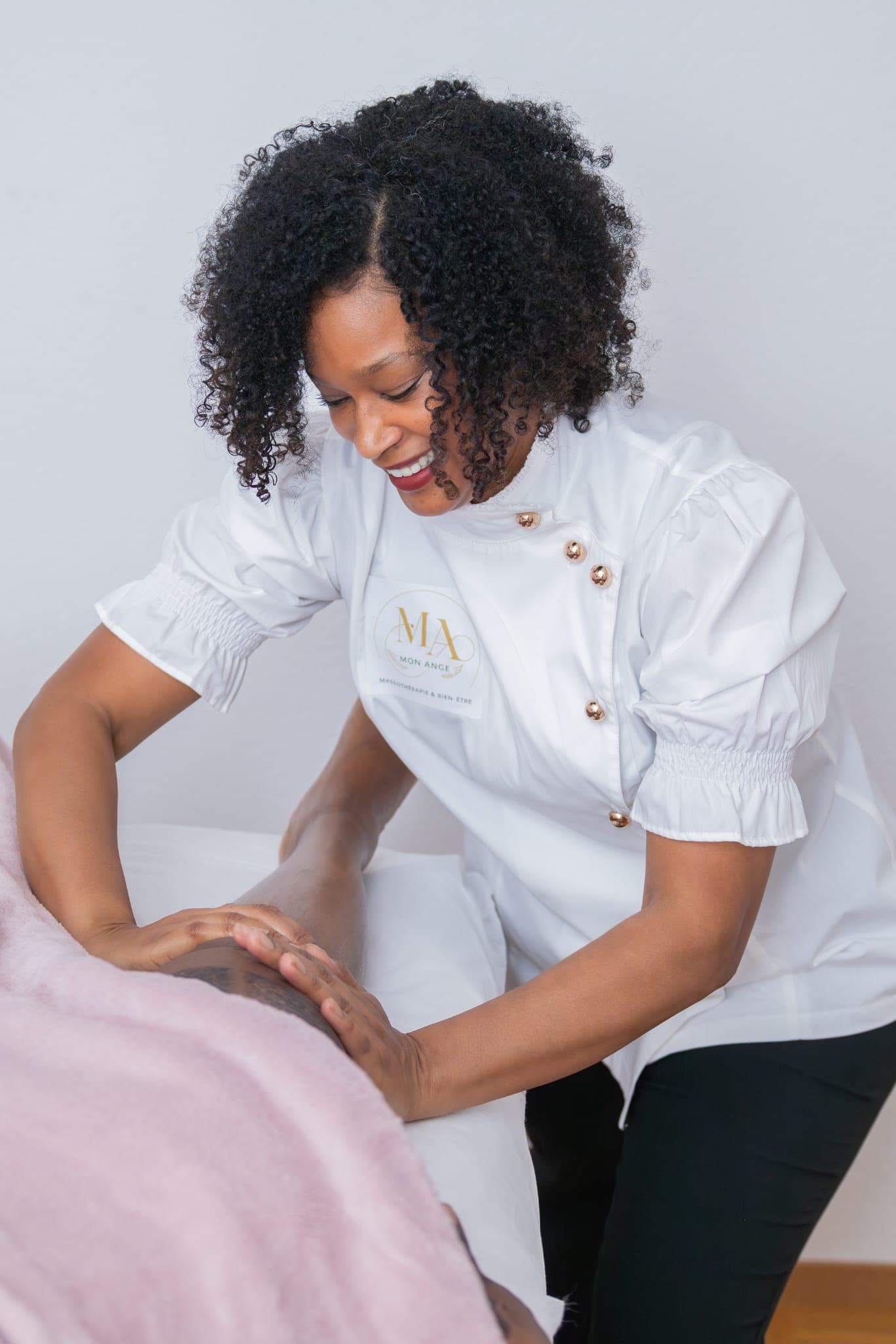 Une massothérapeute souriante en uniforme blanc effectue un massage des jambes sur un client.