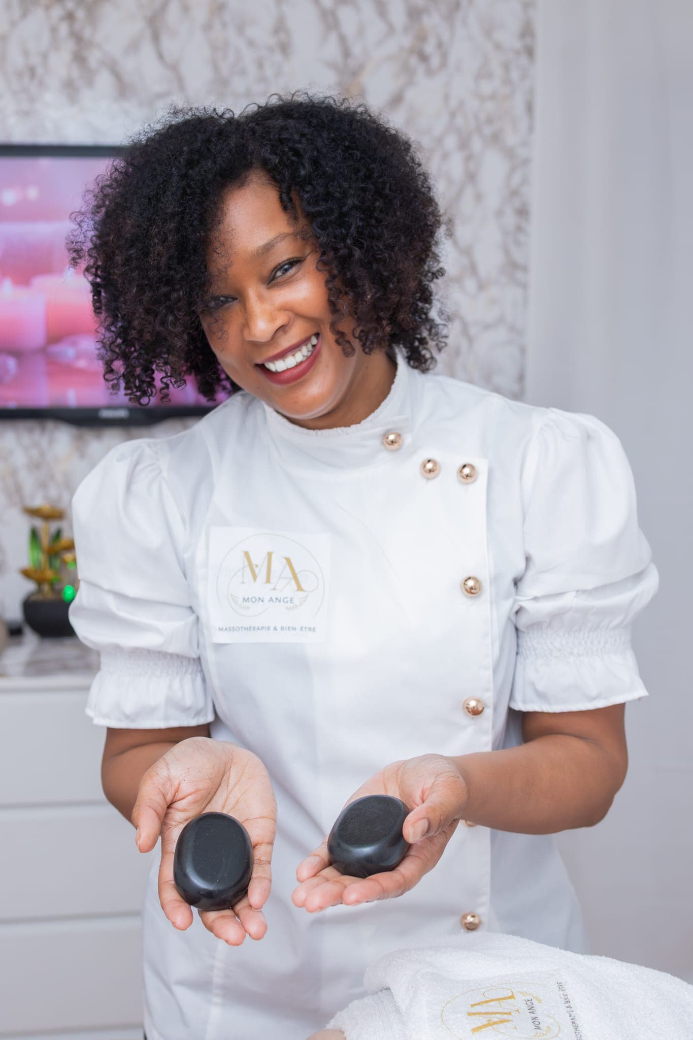 Massothérapeute souriante en uniforme blanc présentant deux pierres de massage noires et lisses.