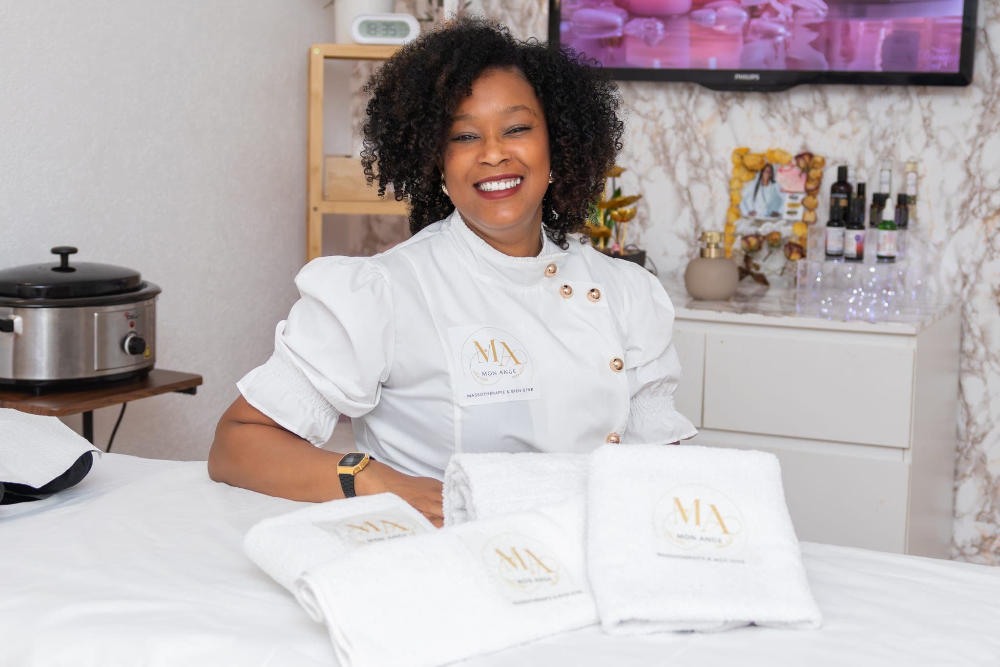 Massothérapeute souriante en uniforme blanc posant derrière des serviettes blanches dans son spa.